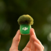 Close-up view of DJI Mic Mini 2 Transmitter held between fingers with furry windscreen attached against blurred green background