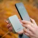Hand holding a sage green Verbatim WPB20 power bank next to a smartphone.