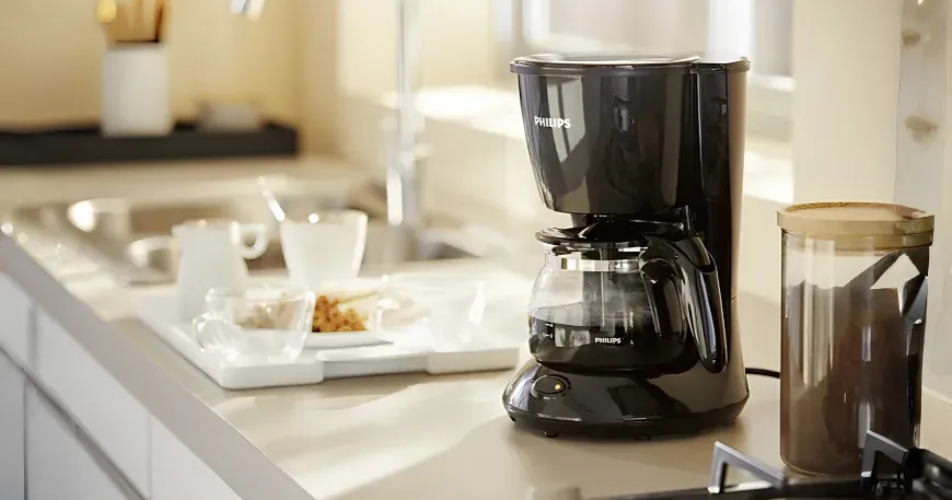 Side-angled view of a classic black Philips HD7432 drip coffee maker on a kitchen counter with a tray of cups.